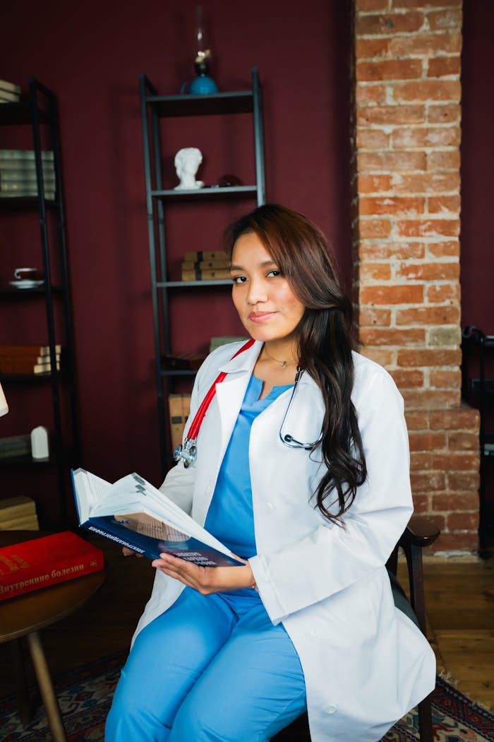 Young female doctor in scrubs reading a medical book in a cozy office setting.